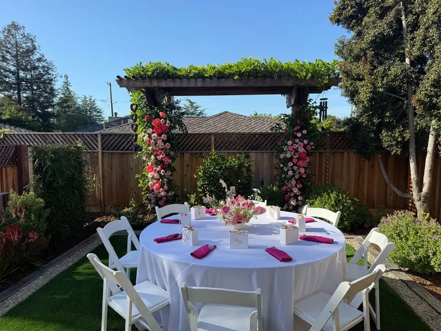 Rose arch in blush and deep red framing a round garden table finished in white linen with fuchsia napkins — floral install styling from our Mountain View catering and florals team